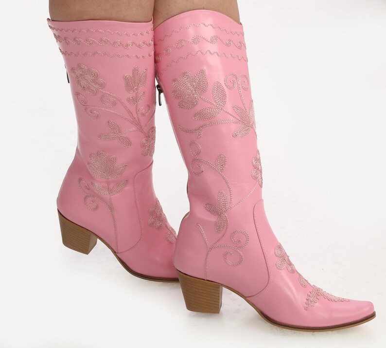 May include: Pink leather cowboy boots with floral embroidery. The boots have a pointed toe and a block heel.