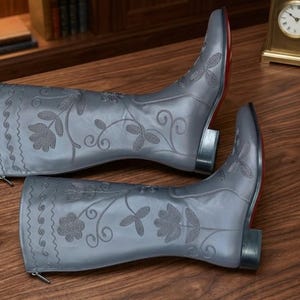 Puede incluir: Un par de botas de vaquero de cuero gris con bordados florales. Las botas tienen punta afilada, tac&oacute;n bajo y suela roja. Las botas miden aproximadamente 30 cm de alto.