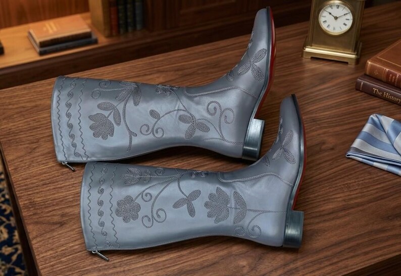 Puede incluir: Un par de botas de vaquero de cuero gris con bordados florales. Las botas tienen punta afilada, tac&oacute;n bajo y suela roja. Las botas miden aproximadamente 30 cm de alto.
