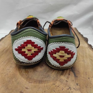K&ouml;nnte beinhalten: Ein Paar gr&uuml;ne Lederschuhe mit einem wei&szlig;en und roten gewebten Design auf der R&uuml;ckseite. Die Schuhe haben eine braune Ledersohle und einen schwarzen Lederbesatz.