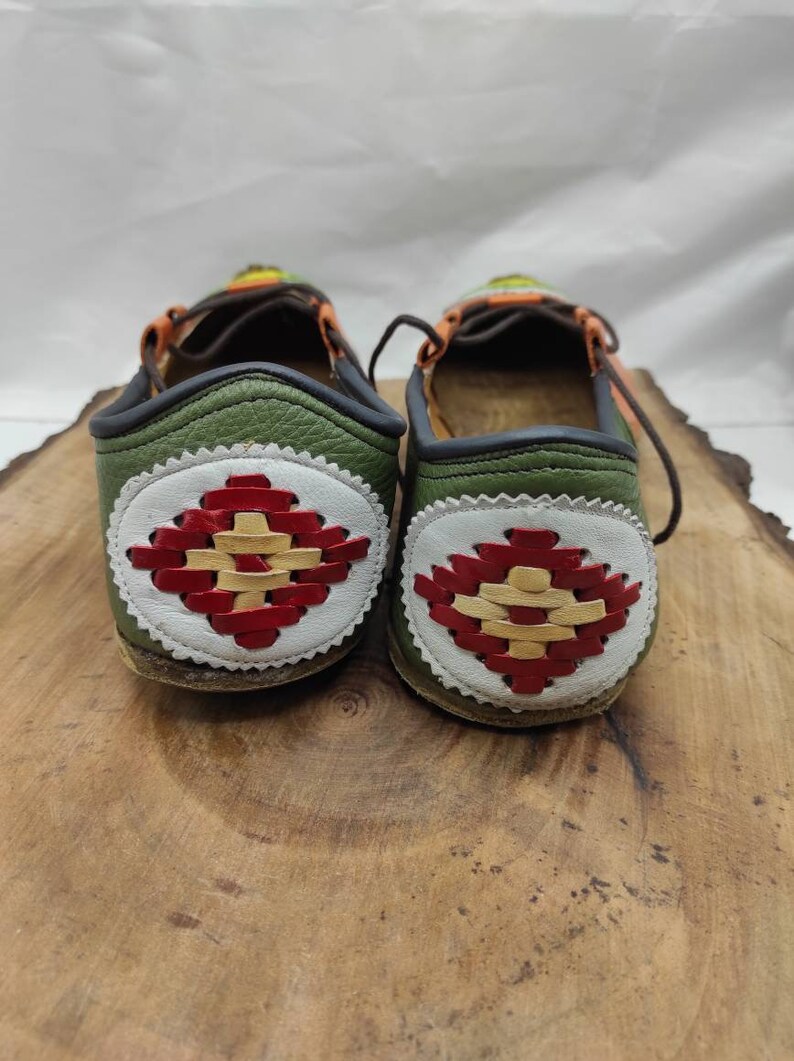 K&ouml;nnte beinhalten: Ein Paar gr&uuml;ne Lederschuhe mit einem wei&szlig;en und roten gewebten Design auf der R&uuml;ckseite. Die Schuhe haben eine braune Ledersohle und einen schwarzen Lederbesatz.