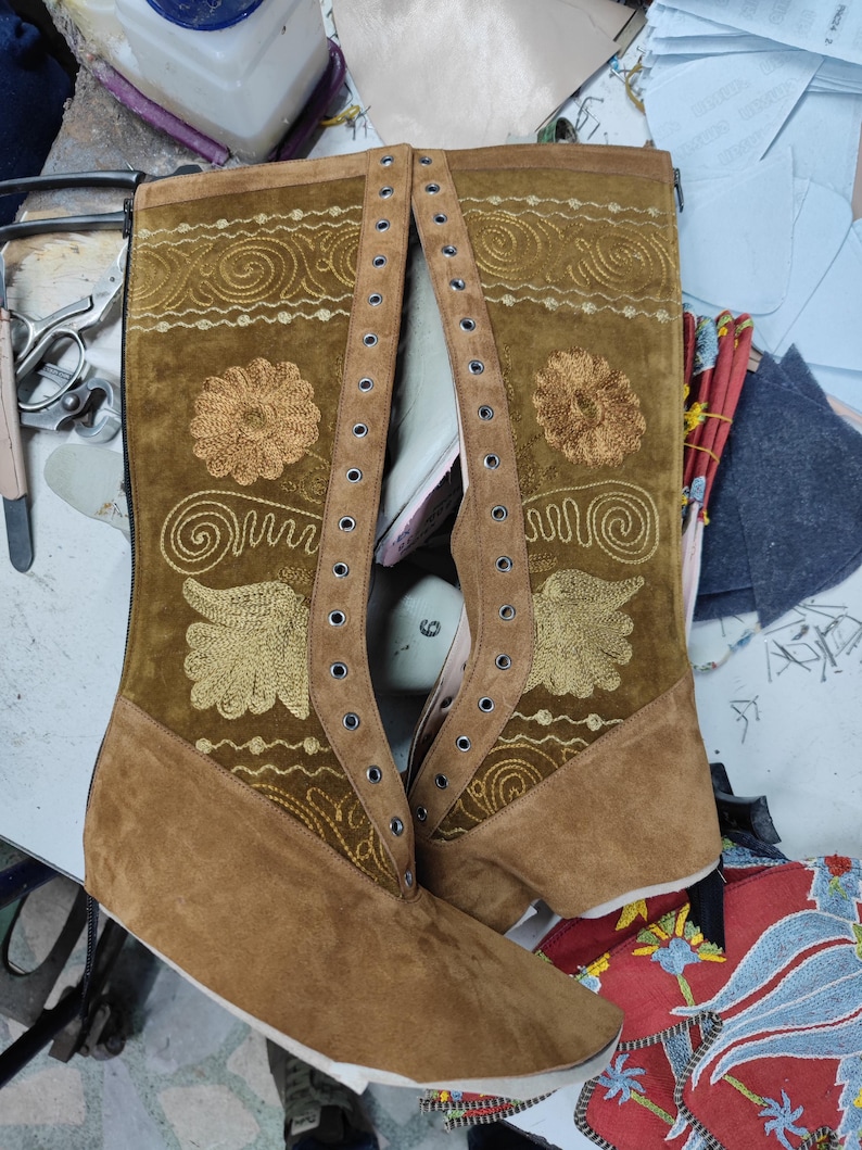 May include: A pair of brown leather boots with gold embroidery and eyelets. The boots have a pointed toe and a low heel.