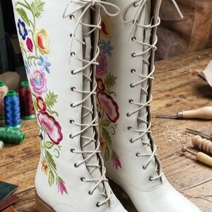 May include: A pair of white leather boots with colorful floral embroidery. The boots have a lace-up design and a low heel. The embroidery features pink, blue, yellow, and green flowers and leaves.