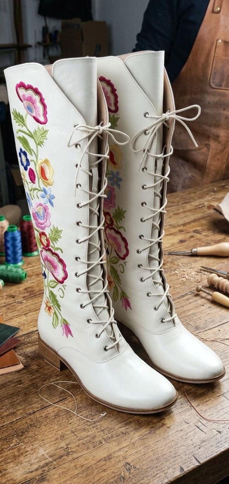 May include: A pair of white leather boots with colorful floral embroidery. The boots have a lace-up design and a low heel. The embroidery features pink, blue, yellow, and green flowers and leaves.