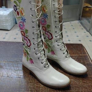 May include: A pair of white knee-high boots with colorful floral embroidery. The boots have a lace-up design and a low heel. The floral embroidery includes red, pink, yellow, and blue flowers.