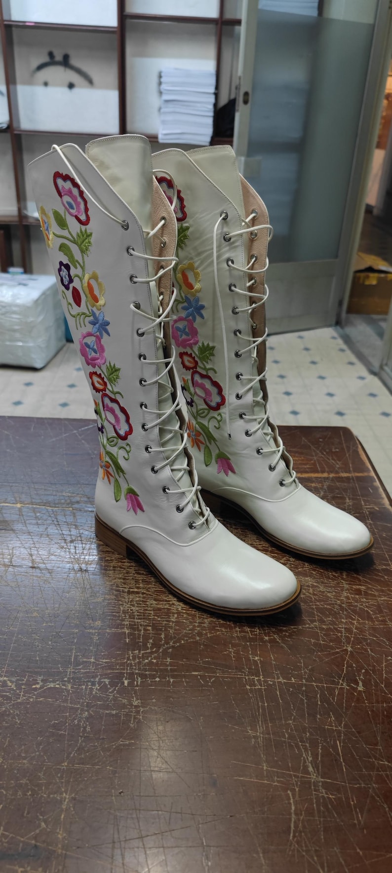 May include: A pair of white knee-high boots with colorful floral embroidery. The boots have a lace-up design and a low heel. The floral embroidery includes red, pink, yellow, and blue flowers.