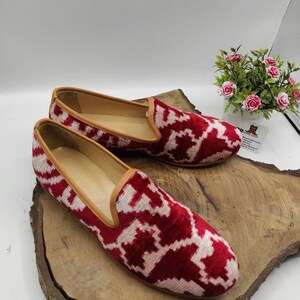 May include: A pair of red and white patterned velvet loafers with a brown leather trim. The loafers are sitting on a wooden surface.
