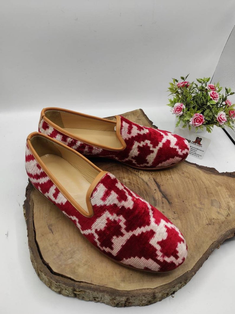 May include: A pair of red and white patterned velvet loafers with a brown leather trim. The loafers are sitting on a wooden surface.