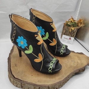 May include: A pair of black leather peep-toe heeled booties with floral embroidery in shades of blue, green, and orange. The booties have a zipper on the back and a decorative chain detail around the ankle.