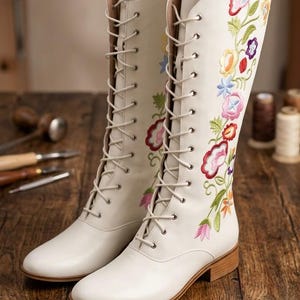 May include: Cream-colored, knee-high boots with floral embroidery. The boots feature a lace-up design and a low heel. Colorful embroidered flowers adorn the sides of the boots, adding a decorative touch. The boots are set on a wooden surface.