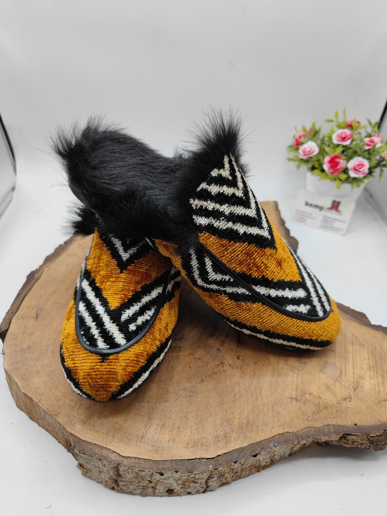 Puede incluir: Un par de zapatillas de terciopelo amarillo mostaza con patrones geom&eacute;tricos en blanco y negro. Las zapatillas tienen un ribete de piel sint&eacute;tica negra alrededor de la parte superior.