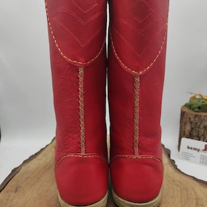 May include: A pair of red leather boots with yellow stitching and a brown sole. The boots are tall and have a pointed toe.