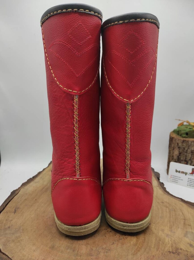 May include: A pair of red leather boots with yellow stitching and a brown sole. The boots are tall and have a pointed toe.