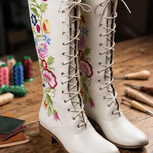May include: A pair of white knee-high boots with colorful floral embroidery. The boots have a lace-up design and a low heel. The embroidery features pink, blue, yellow, and green flowers. The boots are displayed on a wooden surface.