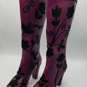 Puede incluir: Un par de botas moradas hasta la rodilla con bordados florales negros y un patr&oacute;n de l&iacute;neas onduladas cerca de la parte superior. Las botas tienen punta redondeada, tac&oacute;n de bloque y cierre de cremallera lateral sobre el cuero morado.
