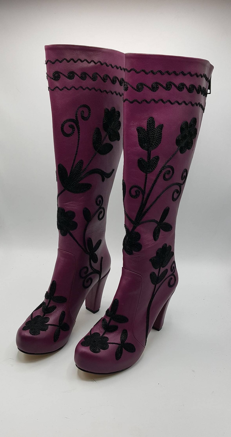 Puede incluir: Un par de botas moradas hasta la rodilla con bordados florales negros y un patr&oacute;n de l&iacute;neas onduladas cerca de la parte superior. Las botas tienen punta redondeada, tac&oacute;n de bloque y cierre de cremallera lateral sobre el cuero morado.