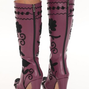 Puede incluir: Un par de botas hasta la rodilla de color morado ciruela con bordados negros de motivos florales y volutas. Las botas tienen tac&oacute;n de bloque, una cremallera vertical completa en la parte posterior y un acabado liso con costuras.