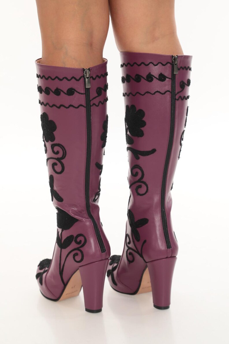 Puede incluir: Un par de botas hasta la rodilla de color morado ciruela con bordados negros de motivos florales y volutas. Las botas tienen tac&oacute;n de bloque, una cremallera vertical completa en la parte posterior y un acabado liso con costuras.