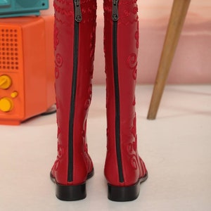 Puede incluir: Un par de botas rojas hasta la rodilla con cremalleras negras en la parte trasera. Las botas tienen patrones bordados decorativos en un color rojo a juego. Las botas tienen un peque&ntilde;o tac&oacute;n de bloque negro.