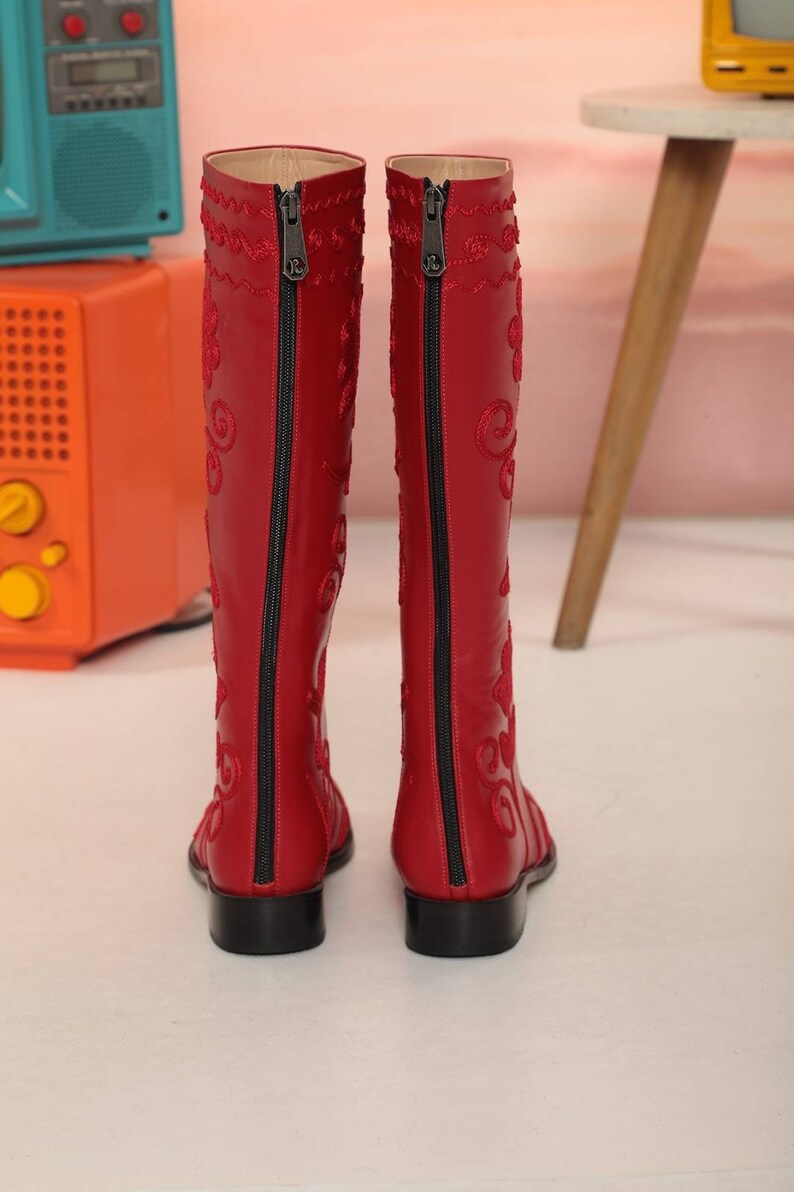 Puede incluir: Un par de botas rojas hasta la rodilla con cremalleras negras en la parte trasera. Las botas tienen patrones bordados decorativos en un color rojo a juego. Las botas tienen un peque&ntilde;o tac&oacute;n de bloque negro.