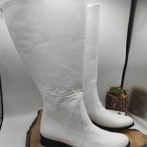 Puede incluir: Un par de botas blancas de cuero de ca&ntilde;a alta con tac&oacute;n bajo. Las botas est&aacute;n sobre una superficie de madera.
