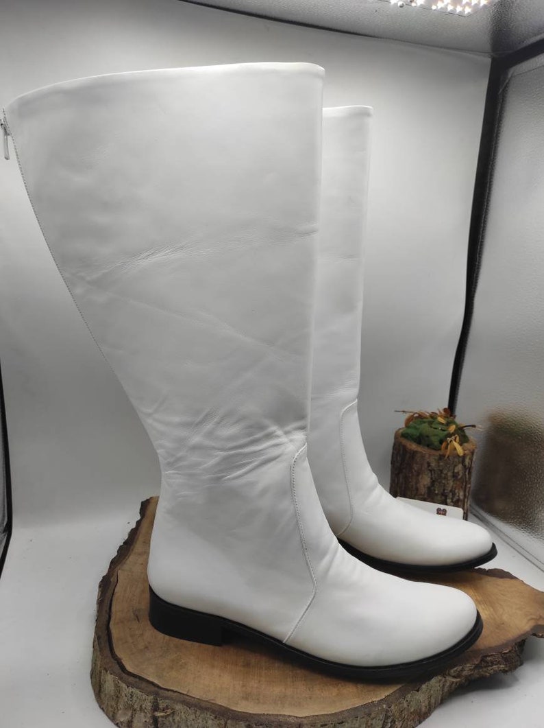 Puede incluir: Un par de botas blancas de cuero de ca&ntilde;a alta con tac&oacute;n bajo. Las botas est&aacute;n sobre una superficie de madera.