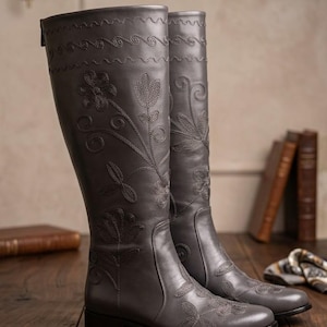 Puede incluir: Un par de botas altas de cuero gris con bordados florales. Las botas presentan costuras decorativas en la parte superior y patrones florales en la parte delantera. Se exhiben sobre una superficie de madera.