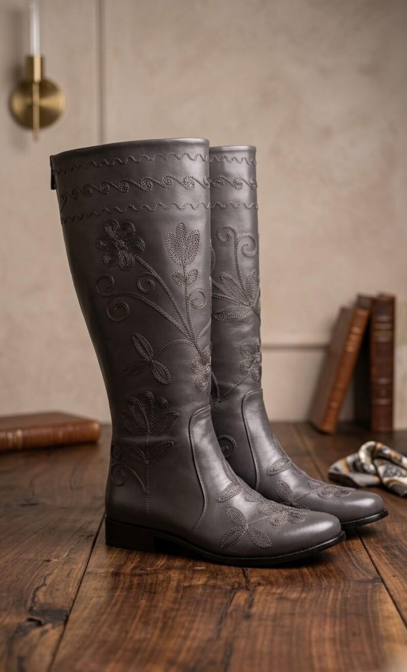 Puede incluir: Un par de botas altas de cuero gris con bordados florales. Las botas presentan costuras decorativas en la parte superior y patrones florales en la parte delantera. Se exhiben sobre una superficie de madera.