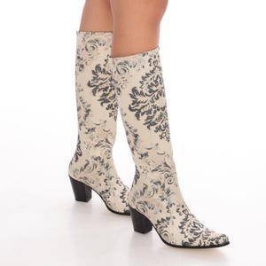 May include: A pair of knee-high boots featuring a cream-colored base with a dark gray damask pattern. The boots have a pointed toe, a structured shaft that reaches the lower calf, and a solid black block heel for stability and support.