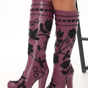 Puede incluir: Botas hasta la rodilla de color morado ciruela intenso con bordados florales negros que se extienden desde la punta redondeada hasta la ca&ntilde;a. Las botas tienen un tac&oacute;n de bloque grueso y una l&iacute;nea decorativa de costura negra ondulada.