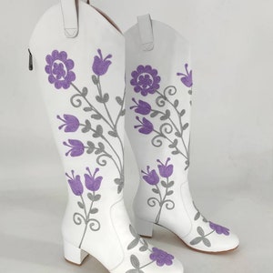 May include: A pair of white leather knee-high boots with a floral embroidery design. The flowers are purple and gray.