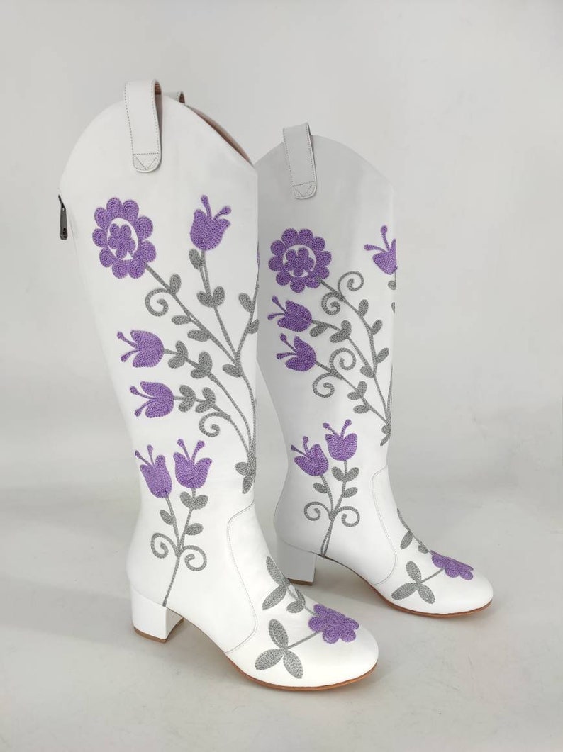 May include: A pair of white leather knee-high boots with a floral embroidery design. The flowers are purple and gray.