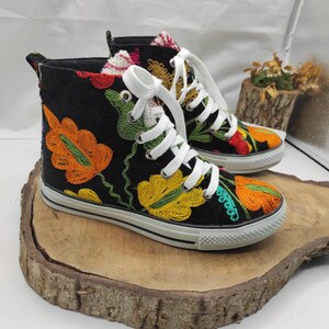 May include: Black high-top canvas shoes with colorful embroidered floral designs. The shoes have white laces and a white rubber sole.
