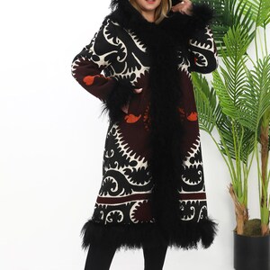 May include: A long black coat with a white and black patterned design. The coat has a hood with a black fur trim. The coat is lined with black fur and has a black fur trim around the cuffs. The coat is being worn by a person with long blonde hair.