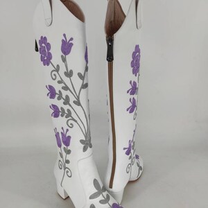 May include: White leather knee-high boots with a block heel and a side zipper. The boots are decorated with purple and gray embroidered flowers.