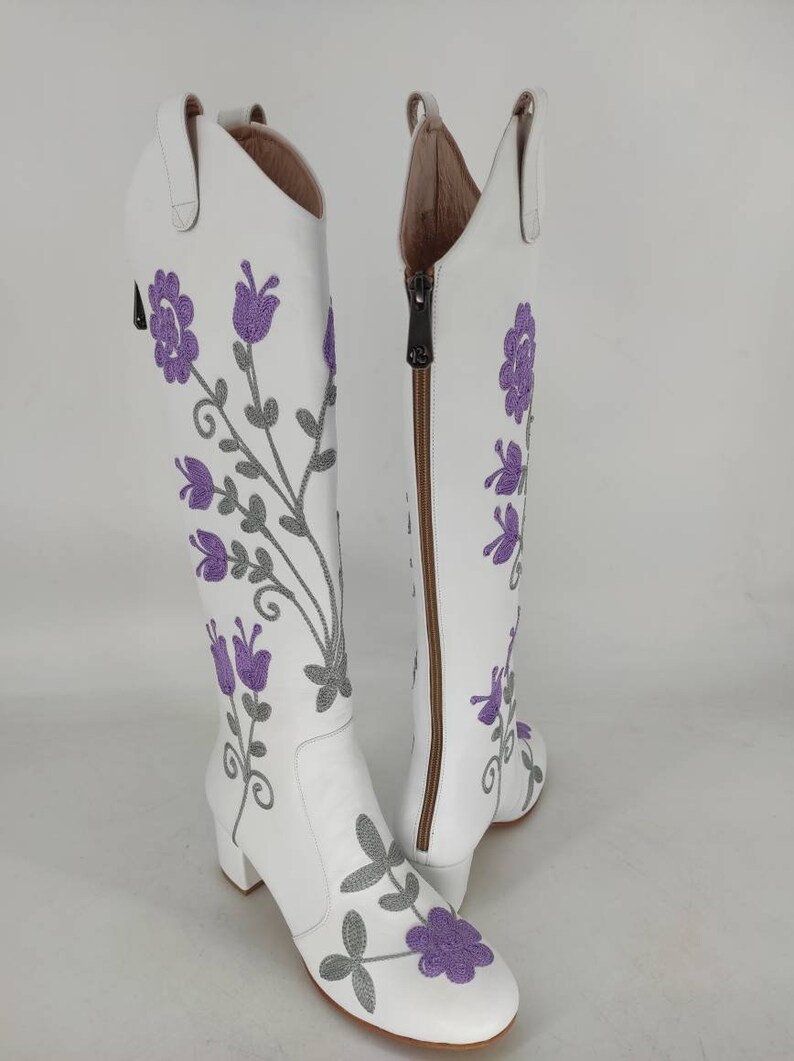 May include: White leather knee-high boots with a block heel and a side zipper. The boots are decorated with purple and gray embroidered flowers.