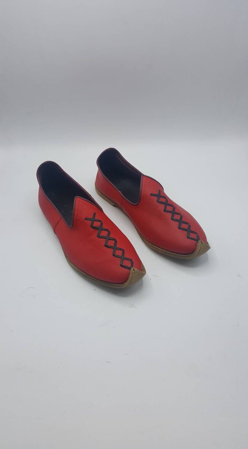 May include: A pair of red leather slip-on shoes with black stitching in a diamond pattern. The shoes have a pointed toe and a brown leather sole.