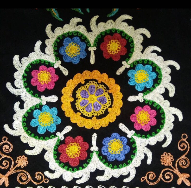 May include: A black fabric with colorful embroidered flowers in a circular pattern. The flowers are red, pink, yellow, blue, and green. The center of the circle has a yellow and purple embroidered design.