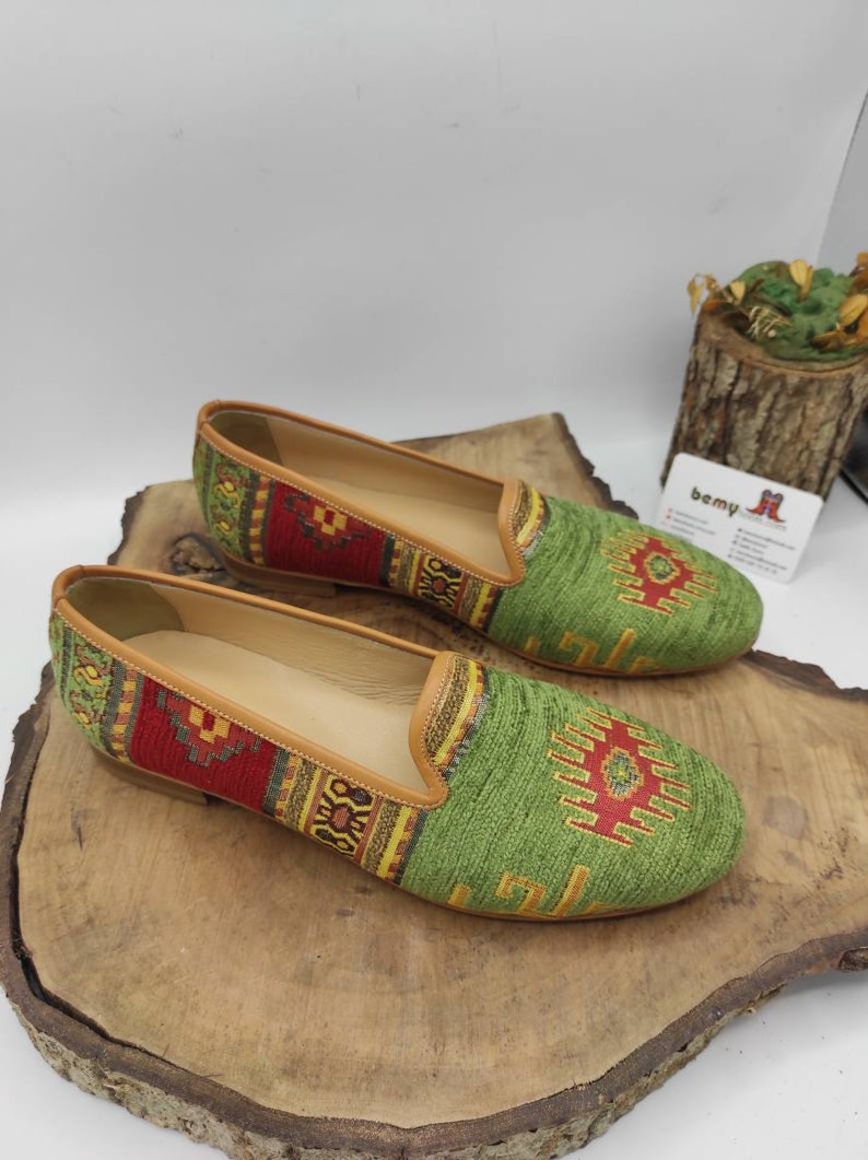 May include: A pair of green and red patterned slip-on shoes with tan leather trim. The shoes have a woven textile design and are displayed on a wooden surface. The shoes have a classic loafer style.