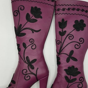 Puede incluir: Un par de botas moradas hasta la rodilla con patrones florales bordados en negro y enredaderas. Las botas tienen punta redondeada, tac&oacute;n de bloque y cierre de cremallera lateral. La parte superior de la ca&ntilde;a presenta un patr&oacute;n de ondas horizontal.