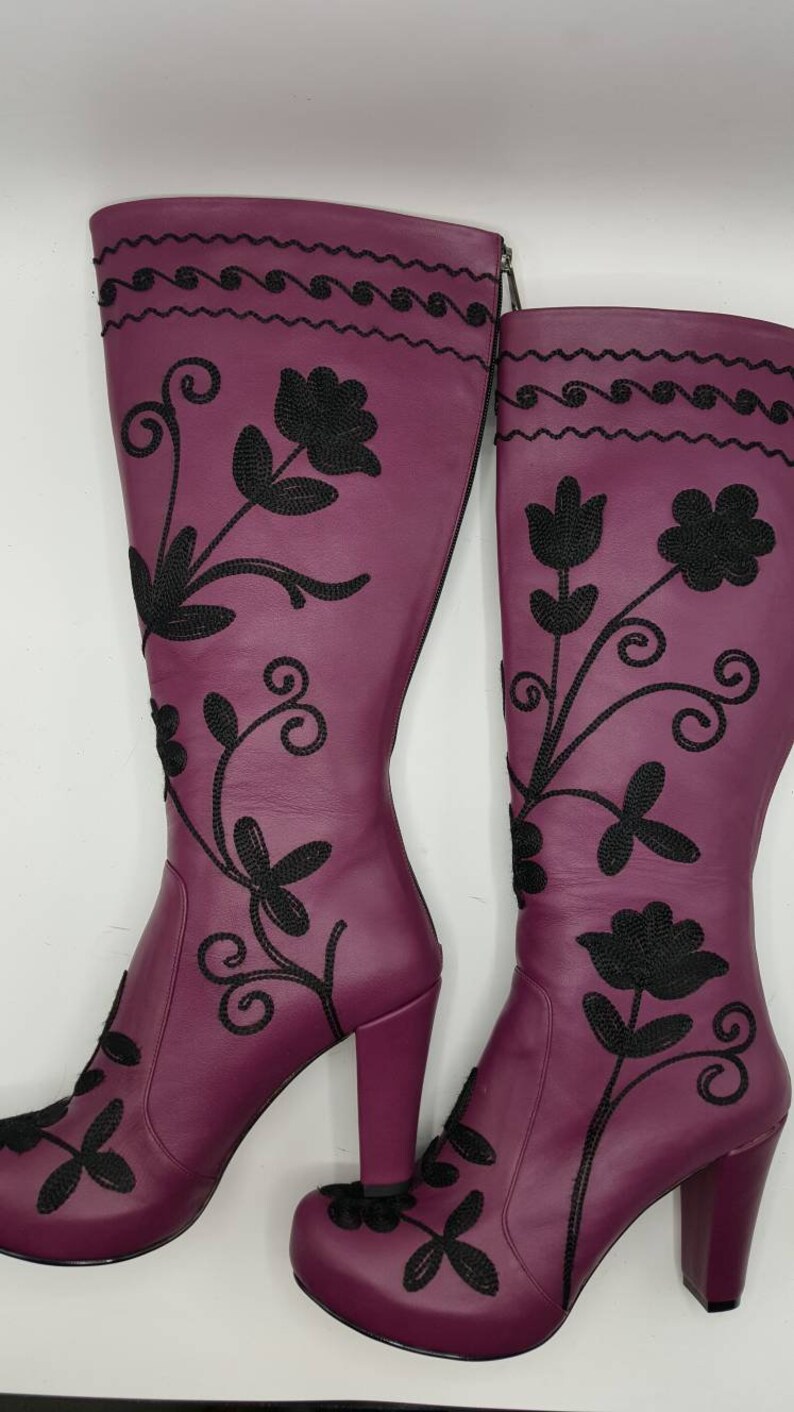 Puede incluir: Un par de botas moradas hasta la rodilla con patrones florales bordados en negro y enredaderas. Las botas tienen punta redondeada, tac&oacute;n de bloque y cierre de cremallera lateral. La parte superior de la ca&ntilde;a presenta un patr&oacute;n de ondas horizontal.