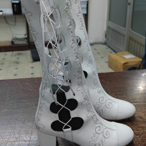 May include: White leather boots with lace-up detailing and floral embroidery. The boots feature a mid-height heel and cut-out patterns. They are displayed on a brown wooden surface.