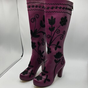 Puede incluir: Un par de botas moradas hasta la rodilla con patrones florales bordados en negro y un dise&ntilde;o de l&iacute;nea ondulada cerca de la parte superior. Las botas tienen tac&oacute;n de bloque, punta redondeada y una cremallera vertical en la parte trasera.