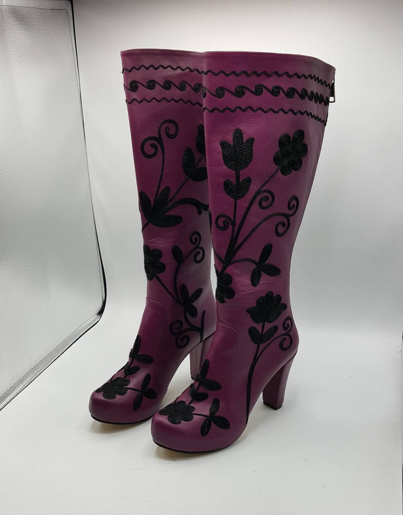 Puede incluir: Un par de botas moradas hasta la rodilla con patrones florales bordados en negro y un dise&ntilde;o de l&iacute;nea ondulada cerca de la parte superior. Las botas tienen tac&oacute;n de bloque, punta redondeada y una cremallera vertical en la parte trasera.