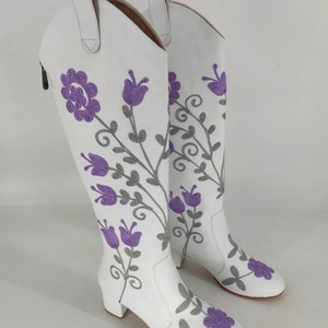 May include: White leather knee-high boots with a floral embroidery design. The flowers are purple and gray.