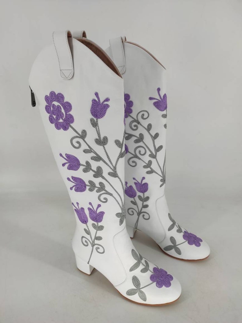 May include: White leather knee-high boots with a floral embroidery design. The flowers are purple and gray.