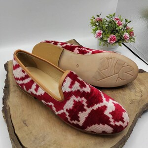 May include: A pair of red and white patterned velvet slippers with a brown leather sole. The slippers are on a wooden surface.