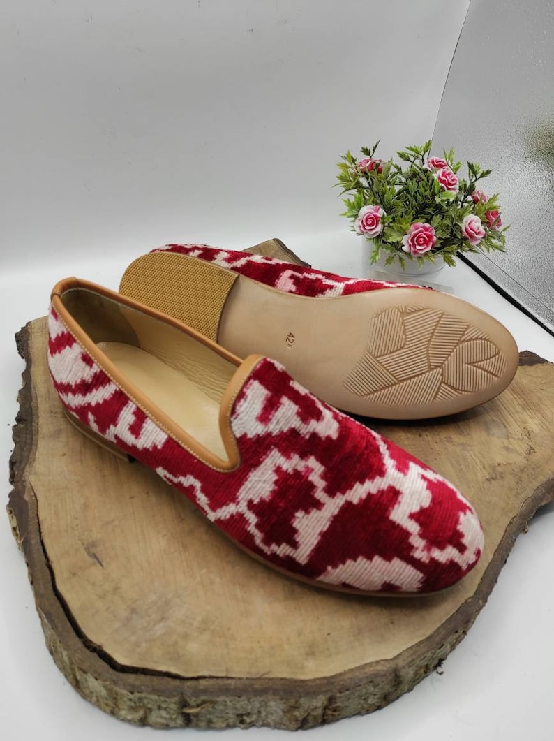 May include: A pair of red and white patterned velvet slippers with a brown leather sole. The slippers are on a wooden surface.
