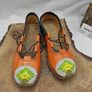 K&ouml;nnte beinhalten: Ein Paar orangefarbene Lederschuhe mit gr&uuml;nen und gelben gewebten Mustern an den Zehen. Die Schuhe haben braune Schn&uuml;rsenkel und stehen auf einer Holzoberfl&auml;che.