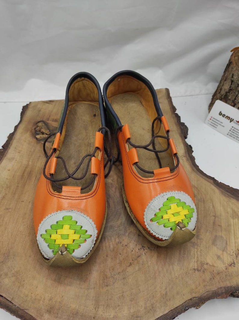 K&ouml;nnte beinhalten: Ein Paar orangefarbene Lederschuhe mit gr&uuml;nen und gelben gewebten Mustern an den Zehen. Die Schuhe haben braune Schn&uuml;rsenkel und stehen auf einer Holzoberfl&auml;che.